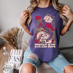 May include: Purple t-shirt with a skull graphic and the text "Staying Alive". The skull is surrounded by flowers and a coffee cup pouring coffee into the skull.