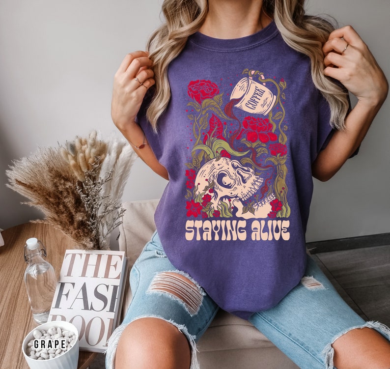 May include: Purple t-shirt with a skull graphic and the text "Staying Alive". The skull is surrounded by flowers and a coffee cup pouring coffee into the skull.