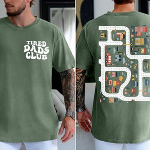 K&ouml;nnte beinhalten: Moosgr&uuml;nes T-Shirt mit dem wei&szlig;en Schriftzug "TIRED DADS CLUB" auf der Vorderseite. Die R&uuml;ckseite zeigt ein Stra&szlig;enkarten-Design mit Autos, Geb&auml;uden und B&auml;umen. Das Shirt ist ein bequemer, l&auml;ssiger Stil.