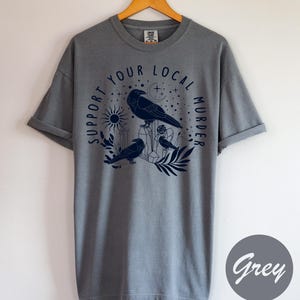 May include: A grey t-shirt with a navy blue graphic featuring three crows, crystals, a sun, and the text "SUPPORT YOUR LOCAL MURDER" in a circular design. The shirt is displayed on a wooden hanger.