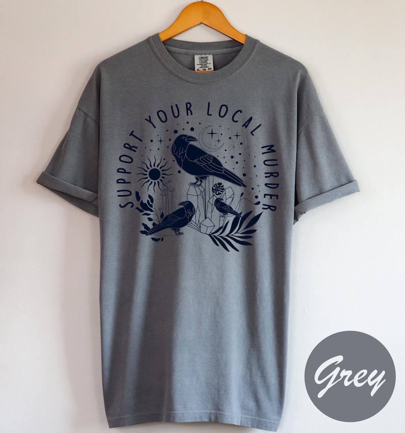 May include: A grey t-shirt with a navy blue graphic featuring three crows, crystals, a sun, and the text "SUPPORT YOUR LOCAL MURDER" in a circular design. The shirt is displayed on a wooden hanger.