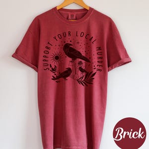 May include: Brick-colored t-shirt with rolled-up sleeves, featuring a black graphic of crows, crystals, and the text "SUPPORT YOUR LOCAL MURDER." The design includes a sun, moon, and stars. The shirt is hanging on a wooden hanger.