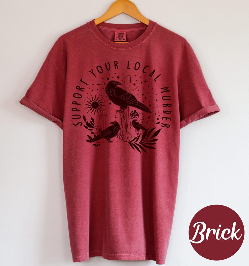 May include: Brick-colored t-shirt with rolled-up sleeves, featuring a black graphic of crows, crystals, and the text "SUPPORT YOUR LOCAL MURDER." The design includes a sun, moon, and stars. The shirt is hanging on a wooden hanger.