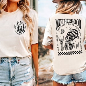 May include: A cream colored t-shirt with a black and white graphic of a skeleton with a butterfly and the text "MOTHERHOOD SOME DAYS I ROCK IT. SOME DAYS IT ROCKS ME. EITHER WAY WE'RE ROCKIN'" on the back. The front of the shirt has a smaller graphic of a skeleton hand with a butterfly and the text "MOTHERHOOD".