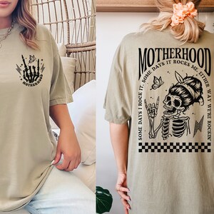 May include: Khaki t-shirt with a black graphic print of a skeleton with a butterfly and the text "Some days I rock it. Some days it rocks me. Either way we're rockin'" on the back. The front of the shirt has a smaller graphic print of a skeleton hand with the text "Motherhood" on the bottom.