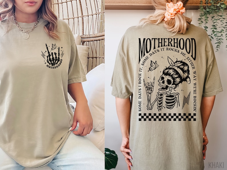 May include: Khaki t-shirt with a black graphic print of a skeleton with a butterfly and the text "Some days I rock it. Some days it rocks me. Either way we're rockin'" on the back. The front of the shirt has a smaller graphic print of a skeleton hand with the text "Motherhood" on the bottom.