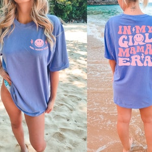 May include: A periwinkle blue short-sleeve t-shirt featuring a small pink smiley face graphic on the front left chest with the text "GIRL MAMA ERA". The back displays a large pink graphic with the text "IN MY GIRL MAMA ERA" and smiley faces.