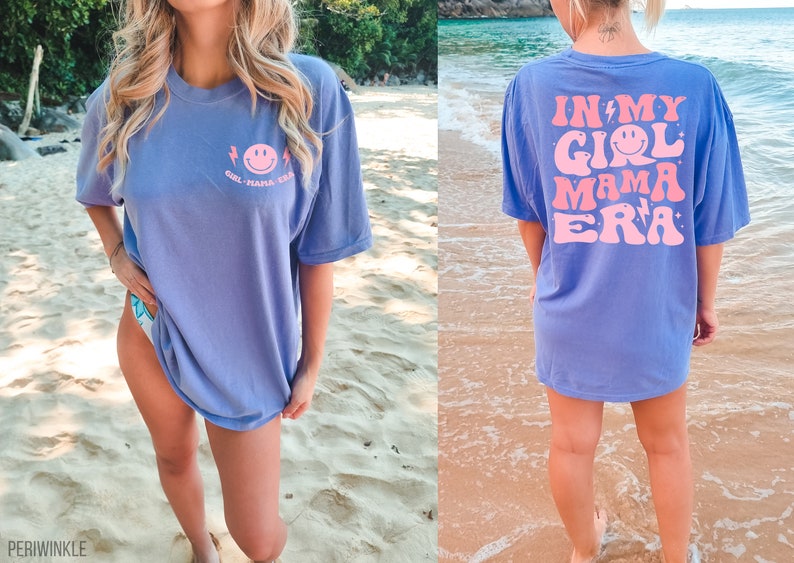 May include: A periwinkle blue short-sleeve t-shirt featuring a small pink smiley face graphic on the front left chest with the text "GIRL MAMA ERA". The back displays a large pink graphic with the text "IN MY GIRL MAMA ERA" and smiley faces.