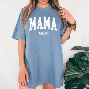 May include: A light blue t-shirt with the word "MAMA" printed in white with a distressed look. Below the word is a white oval with the text "EST. 2004" inside.