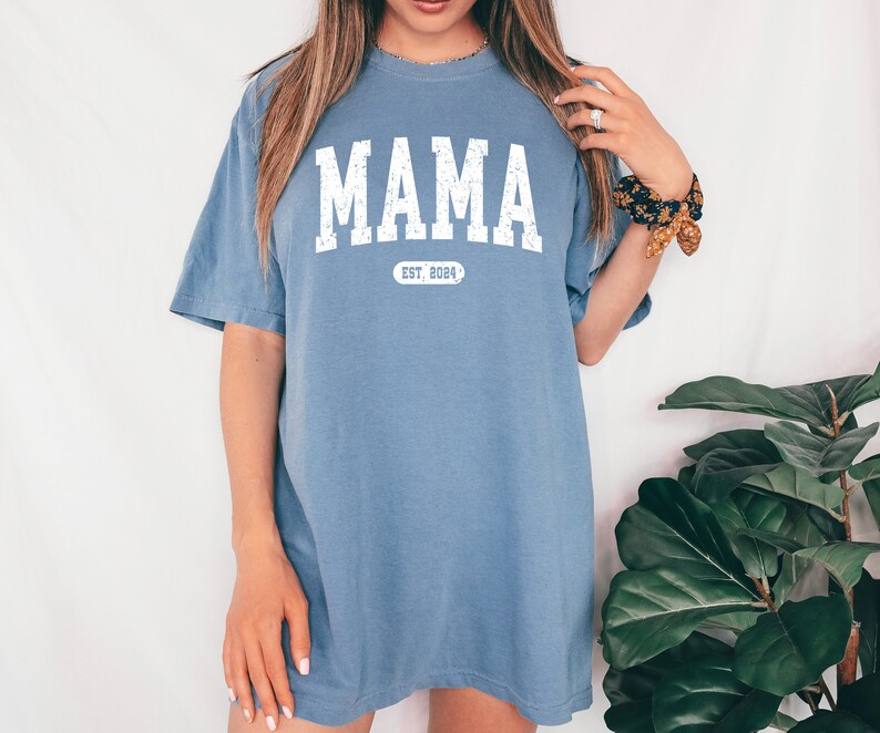 May include: A light blue t-shirt with the word "MAMA" printed in white with a distressed look. Below the word is a white oval with the text "EST. 2004" inside.