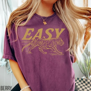 May include: Berry-colored t-shirt with the word "EASY" in gold lettering above a gold tiger graphic. The tiger is walking to the right. The shirt is a crew neck and has short sleeves.