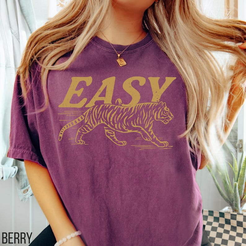 May include: Berry-colored t-shirt with the word "EASY" in gold lettering above a gold tiger graphic. The tiger is walking to the right. The shirt is a crew neck and has short sleeves.