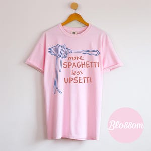 May include: Pink t-shirt with a blue and orange illustration of spaghetti on a fork and the text "more spaghetti less upsetti".