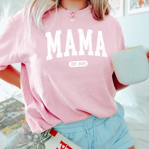 May include: Pink t-shirt with the word "MAMA" in white with a distressed font and the text "EST. 2024" underneath.