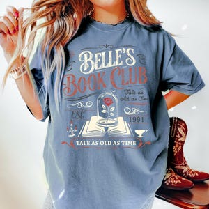 May include: Blue jean-colored t-shirt with the text "BELLE'S BOOK CLUB" in white and red. The design includes a rose in a glass dome, an open book, and the phrase "TALE AS OLD AS TIME". The shirt also has the year "1991" printed on it.