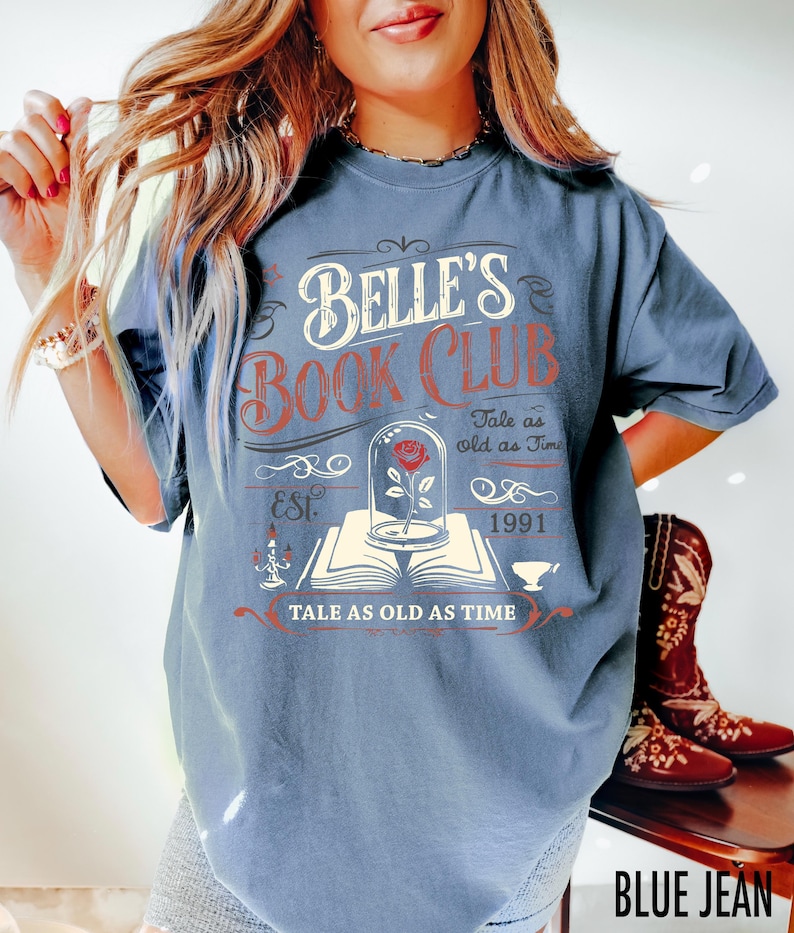 May include: Blue jean-colored t-shirt with the text "BELLE'S BOOK CLUB" in white and red. The design includes a rose in a glass dome, an open book, and the phrase "TALE AS OLD AS TIME". The shirt also has the year "1991" printed on it.