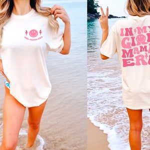May include: An ivory short-sleeve t-shirt featuring a small pink smiley face and lightning bolt graphic on the front chest with the text "GIRL MAMA ERA". The back displays a large pink graphic with the text "IN MY GIRL MAMA ERA" and stars.