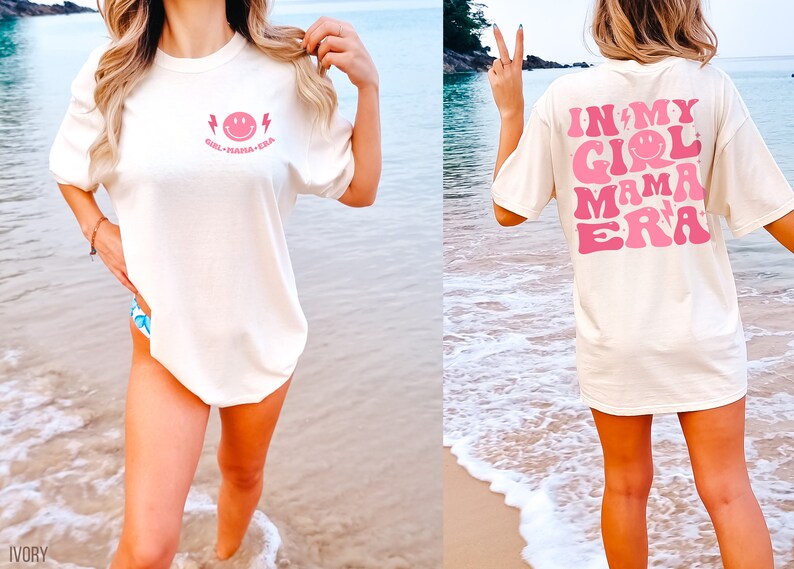 May include: An ivory short-sleeve t-shirt featuring a small pink smiley face and lightning bolt graphic on the front chest with the text "GIRL MAMA ERA". The back displays a large pink graphic with the text "IN MY GIRL MAMA ERA" and stars.