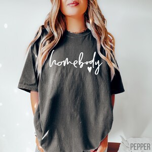 Puede incluir: Una camiseta gris oscuro con el texto "homebody" en letras blancas y dos peque&ntilde;os corazones blancos.