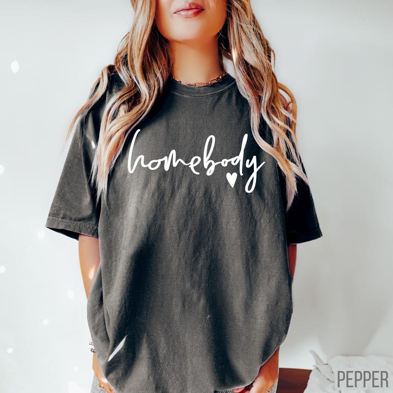 Puede incluir: Una camiseta gris oscuro con el texto "homebody" en letras blancas y dos peque&ntilde;os corazones blancos.
