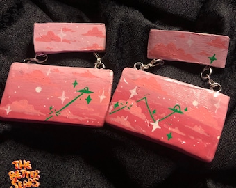 May include: Two pairs of pink painted wooden earrings with a pink and white sky scene with green UFOs and stars. The earrings are attached to silver chains.