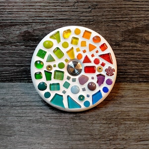Könnte beinhalten: Ein runder, weißer Mosaik-Untersetzer mit einem Regenbogen aus farbigen Glasstücken. Der Untersetzer hat einen zentralen, silberfarbenen Knopf. Das Mosaik umfasst verschiedene Formen und Größen von Glas und schafft so einen lebendigen und dekorativen Artikel.