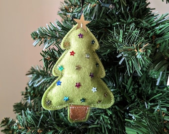 Può includere: Un ornamento per albero di Natale a forma di albero di Natale in feltro verde con un tronco in feltro marrone e una stella dorata in cima. L'ornamento è decorato con paillettes colorate.