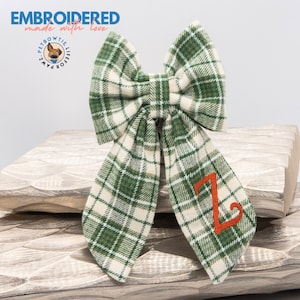 May include: Green and white plaid bow tie with the letter "Z" embroidered in orange thread. The bow tie is made of fabric and is tied with a knot. The bow tie is on a wooden surface.