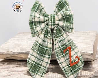 Op de afbeelding: Groen en wit geruit strikje met de letter "Z" geborduurd in oranje draad. Het strikje is gemaakt van stof en is met een knoop vastgemaakt. Het strikje ligt op een houten oppervlak.
