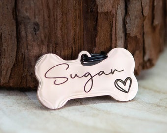 May include: A pink bone-shaped dog tag with the name "Sugar" engraved in cursive script and a small heart symbol.