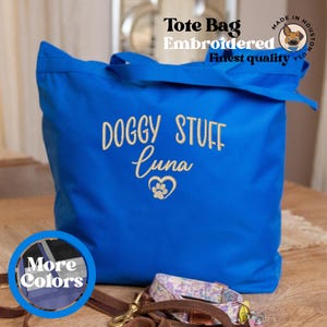 Op de afbeelding: Blauwe tote bag met de tekst "Doggy Stuff Luna" geborduurd in wit. De tas heeft een hart met een pootprint erin. De tas ligt op een houten oppervlak met een bruine leren riem en een roze en witte gepatenteerde riem.