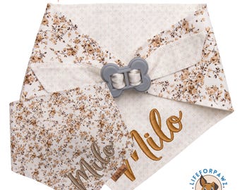 May include: A dog bandana and face mask set in cream with a brown floral pattern. The bandana is personalised with the name "Milo" in brown embroidery. The text "EMBROIDERED made with love" is at the top. Includes a grey bone-shaped clip.