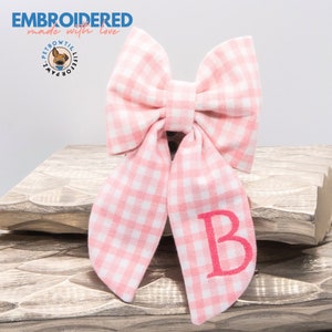 May include: Pink and white gingham bow tie with the letter "B" embroidered in pink. The bow tie is on a wooden surface. The text "EMBROIDERED made with love" is at the top of the image. The text "PETBOWTIE LIFE FOR PAWZ" is in a circle with a dog illustration.