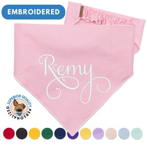 May include: Pink dog bandana with white embroidered text "Remy". The bandana has a white border and is made of a soft, comfortable fabric. The bandana is available in a variety of colors.