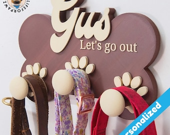 May include: A brown wooden dog bone-shaped wall hook with the name "Gus" and the text "Let's go out" in white lettering. The hook has four white paw print-shaped hooks with three leashes hanging from them.