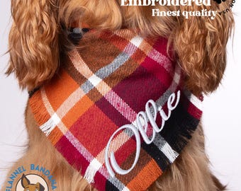 May include: A red, orange, and brown plaid flannel dog bandana with white frayed edges. The bandana is embroidered with the name "Ollie" in white thread. The bandana is worn by a dog with long, brown fur.