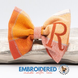 May include: A plaid bow tie in shades of orange, red, and cream, with an embroidered letter "R" in a matching orange thread. The bow tie is made of a textured fabric and is designed for pets. The text "EMBROIDERED made with love" is at the bottom.