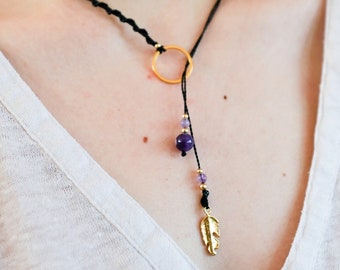 May include: A black cord necklace with a gold circle pendant and a purple gemstone charm. The charm is attached to a gold feather pendant.