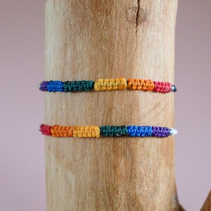 Op de afbeelding: Twee regenboogkleurige armbanden gemaakt van geweven touw met zilveren kralen op een houten achtergrond.