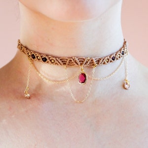 Könnte beinhalten: Eine braune und goldene Choker-Kette mit einem roten Edelstein-Anhänger und kleinen goldenen Charms. Die Kette ist mit zarten goldenen Ketten geschichtet.