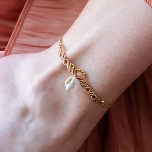 May include: A gold anklet with a small white seashell charm. The anklet is on a person's ankle, and the background is a peach-coloured fabric.