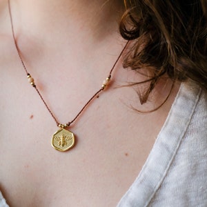 May include: A gold-colored pendant necklace featuring a bee design. The pendant is hexagonal and hangs from a brown cord with small gold beads. The necklace is worn against a light-colored top.