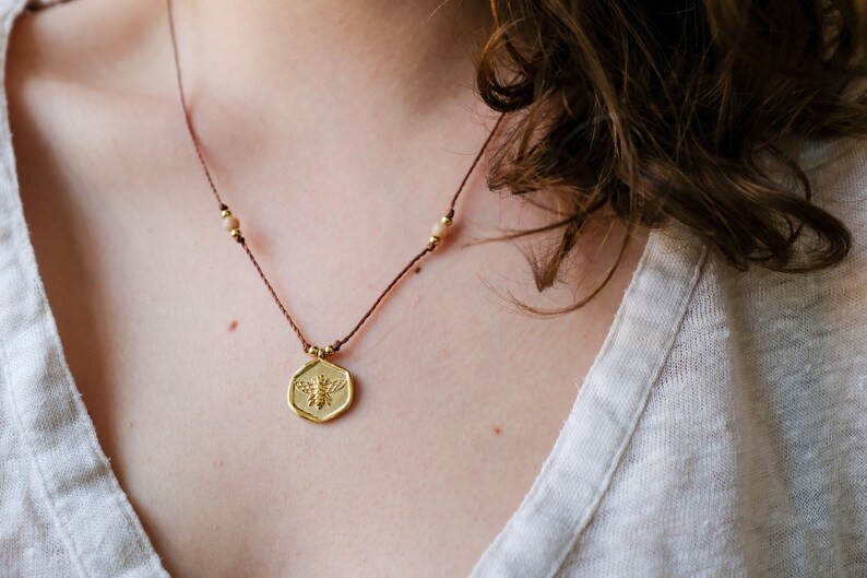 May include: A gold-colored pendant necklace featuring a bee design. The pendant is hexagonal and hangs from a brown cord with small gold beads. The necklace is worn against a light-colored top.