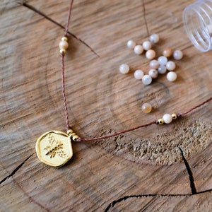 May include: A gold-colored pendant necklace with a bee design, strung on a brown cord. The necklace is adorned with small, round, light-colored beads and gold accents. Scattered around are small, light-colored beads, and a clear plastic container.