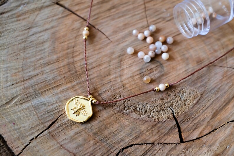May include: A gold-colored pendant necklace with a bee design, strung on a brown cord. The necklace is adorned with small, round, light-colored beads and gold accents. Scattered around are small, light-colored beads, and a clear plastic container.