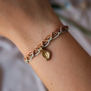 Könnte beinhalten: Ein geflochtenes Armband in Hellbraun und Hellblau mit einem goldenen Blattanhänger.