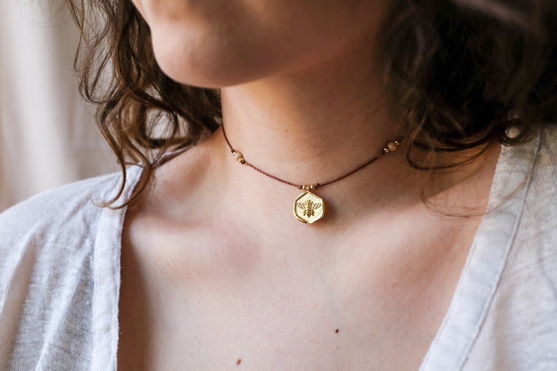 May include: A gold-colored necklace with a bee pendant. The pendant is a hexagon shape with a bee design in the center. The necklace has a brown cord and small beads. The necklace is worn around the neck.