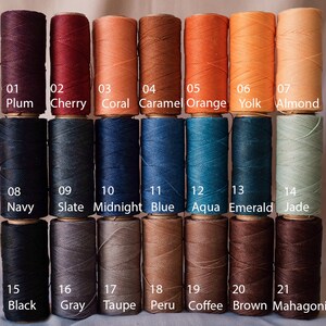May include: A collection of 21 spools of waxed thread in various colors, including plum, cherry, coral, and black. Each spool is labeled with a number and color name. The thread is tightly wound on wooden spools.