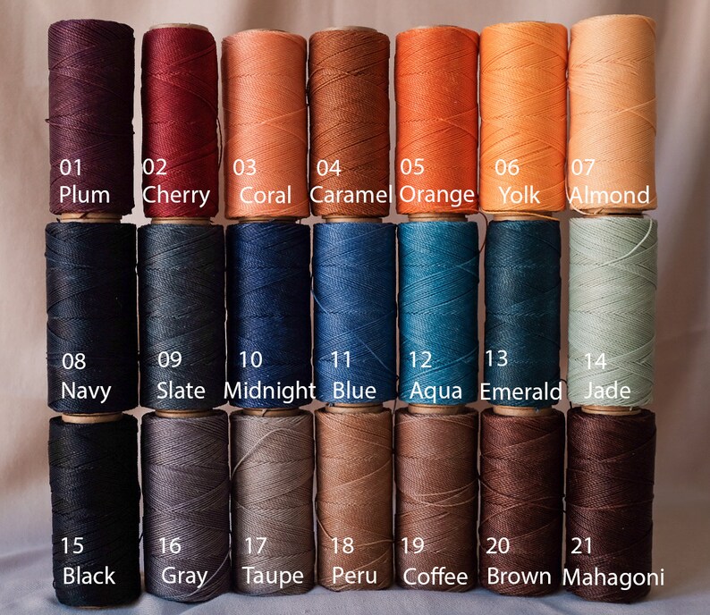 May include: A collection of 21 spools of waxed thread in various colors, including plum, cherry, coral, and black. Each spool is labeled with a number and color name. The thread is tightly wound on wooden spools.