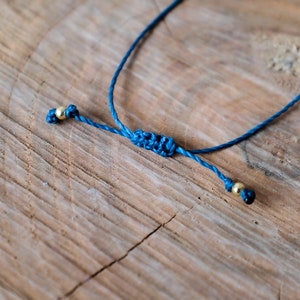 May include: A close-up of a blue adjustable bracelet with gold-colored beads. The bracelet is knotted in the center and rests on a textured wooden surface.